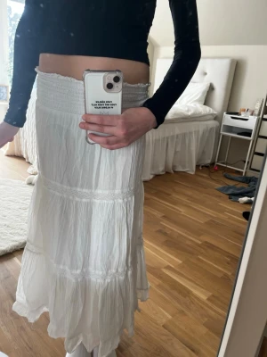 Vit långkjol med volangdetaljer - Säljer en flowy vit långkjol med bred resår i midjan och flera volanglager. Kjolen har en lätt och luftig känsla, perfekt för varmare dagar. Passar till crop tops och sneakers för en chill look.