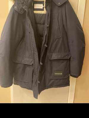 Mörkblå parkas från Calvin Klein Jeans - Snygg mörkblå parkas från Calvin Klein Jeans med stora fickor framtill, dragkedja och knappar. Jackan har en hög krage och huva, perfekt för kalla dagar. Materialet är slitstarkt och jackan har en klassisk, rak passform.