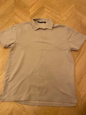 Beige ribbad polotopp med krage - Snygg beige polotopp med korta ärmar och klassisk krage. Toppen har en ribbad struktur som ger en modern känsla och är perfekt för en clean och stilren look. Passar till både jeans och kjol.