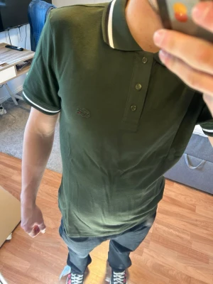 Grön pikétröja från Lacoste - Snygg mörkgrön pikétröja från Lacoste med klassisk krage och vita ränder på krage och ärmslut. Tröjan har tre knappar framtill och den ikoniska krokodilloggan på bröstet. Perfekt för en clean och sportig stil.