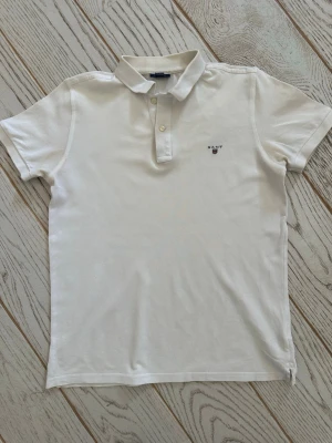 Vit piké t-shirt från Gant - Klassisk vit piké t-shirt från Gant med korta ärmar och liten broderad logga på bröstet. Tröjan har krage och knappar framtill för en stilren look. Tillverkad i mjuk bomull som känns skön mot huden.