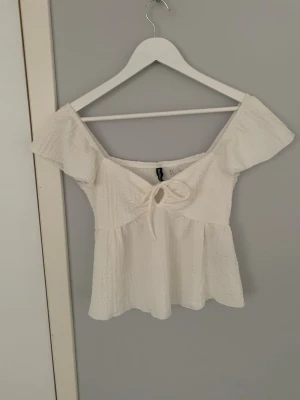 Vit off shoulder topp med knyt - Söt vit topp med off shoulder-modell och korta ärmar. Toppen har en knytning framtill och är gjord i ett lätt, krinklat tyg som ger en avslappnad känsla. Perfekt till sommaren och passar till både jeans och kjol. Köpt på h&m i Spanien och använd en gång.