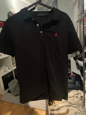 Svart pikétröja från Polo Ralph Lauren - Klassisk svart pikétröja från Polo Ralph Lauren med korta ärmar och krage. Tröjan har knappar framtill och den ikoniska rosa logotypen broderad på bröstet. Tillverkad i mjuk bomull för en bekväm känsla. Helt ny oanvänd!