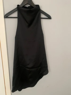 Svart satinklänning med hög hals - Stilren svart klänning i satin med hög hals och ärmlös design. Klänningen har en asymmetrisk nederkant och slät, glansig yta som ger en elegant look. Perfekt för dig som gillar minimalistisk och trendig stil. Öppen i ryggen, aldrig använd.