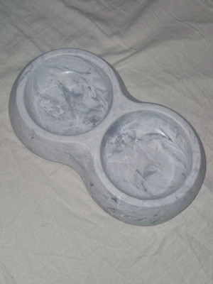 Matskål - Säljer en snygg dubbel matskål i plast med marmorliknande mönster i grått och vitt. Perfekt för husdjur som behöver både mat och vatten på samma plats. Skålen har rundade former och är lätt att rengöra. Bild 1 är ovansidan och bild 2 är undersidan.
