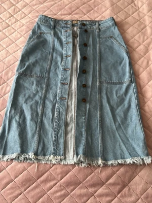 Ljusblå jeanskjol med knappar - Säljer en ljusblå jeanskjol i lång modell med fransad nederkant och knappar framtill. Kjolen har stora fickor på sidorna och bälteshällor i midjan. Perfekt för dig som gillar en avslappnad och trendig stil.