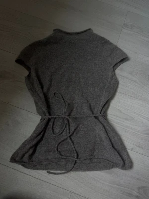 Grå stickad topp med knytband - Mysig grå stickad topp med korta ärmar och knytband i midjan. Modellen har en högre halsringning och är lätt figursydd för en snygg siluett. Perfekt att styla över en långärmad tröja eller bära som den är. Materialet känns mjukt och följsamt.
