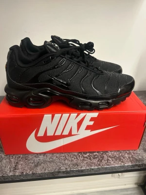 Nike Air Max Plus helsvarta sneakers - Säljer ett par helsvarta Nike Air Max Plus sneakers med mesh- och syntetmaterial, ikoniska vågiga linjer och synlig Air-dämpning i sulan. Skorna har svarta snören och diskreta loggor på plös och häl. Perfekta för dig som gillar streetwear och sportig stil.