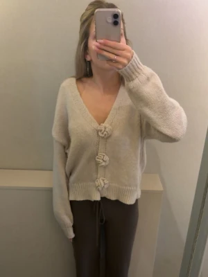 Beige kofta med knytknappar - Mysig beige kofta med v-ringning och tre dekorativa bollknappar framtill. Koftan har en avslappnad passform, långa ärmar och ribbade muddar. Perfekt för lager på lager och enkel att styla till olika outfits. Använd väldigt lite kanske fyra gånger