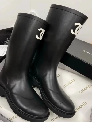 Svarta gummistövlar från Chanel - Snygga svarta gummistövlar från Chanel med det klassiska vita CC-logotypen framtill. Stövlarna har en hög, rak siluett och grov sula med Chanel-logga undertill. Tillverkade i slitstarkt gummi, perfekta för regniga dagar och streetstyle.