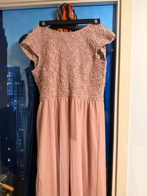 Lång rosa balklänning med snörning i ryggen - Elegant balklänning i ljusrosa med spetsdetaljer upptill och slät kjol. Klänningen har korta ärmar, v-ringad rygg med snörning och ett romantiskt uttryck. Perfekt för speciella tillfällen där du vill känna dig extra fin. Klänningen är stl XL