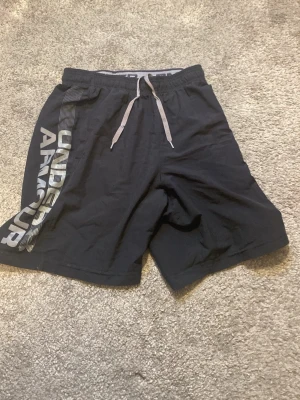 Svarta shorts från Under Armour - Snygga svarta shorts från Under Armour med grå snörning i midjan och stort logotryck längs ena benet. Perfekta för träning eller chill. Lätta och bekväma med normal passform.