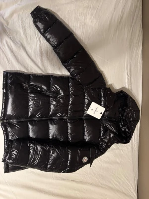 Svart dunjacka från Moncler - Säljer en svart, glansig dunjacka från Moncler med avtagbar huva och två dragkedjeförsedda fickor framtill. Jackan har klassisk puffer-design och Moncler-logga på ärmen. Perfekt för kalla vinterdagar och riktigt snygg att bära utomhus.