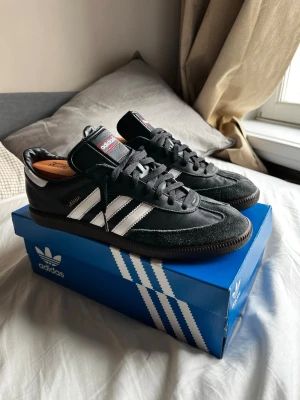 Adidas Samba OG svart/vit sneakers - Klassiska Adidas Samba OG sneakers i svart läder med vita detaljer och bruna gummisulor. Skorna har de ikoniska tre ränderna på sidan, guldfärgad Samba-text och svart plös med Adidas-logga. Perfekt för dig som gillar retrostil och streetwear. LÅDAN INGÅR INTE!
