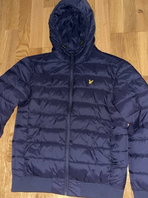 Mörkblå pufferjacka från Lyle & Scott - Snygg mörkblå pufferjacka från Lyle & Scott med gul logga på bröstet. Jackan har huva, dragkedja framtill och ribbade muddar vid ärmslut och nederkant. Perfekt för kalla dagar och ger en sportig look.
