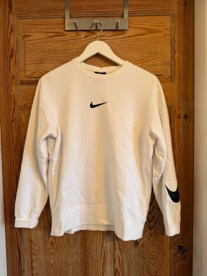Vit tröja från Nike - Säljer en stilren vit sweatshirt från Nike med svart Swoosh-logga på bröstet och ärmen. Tröjan har rund halsringning och långa ärmar. Perfekt för en avslappnad och sportig look.