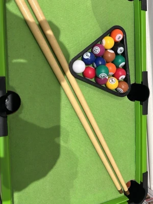 Mini biljardbord med tillbehör - Säljer ett mini biljardbord i grön och svart design med två träköer, en uppsättning färgglada biljardbollar och en svart triangel. Perfekt för spel hemma eller på kontoret. Bordet har klassiska hål och är lätt att flytta.