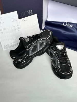 Svarta Dior sneakers med vita detaljer - Snygga svarta Dior sneakers med vita och grå detaljer samt reflekterande inslag. Skorna har en chunky sula, mesh- och läderpaneler samt tydlig Dior-logga på sidan och tungan. Perfekta för dig som vill ha en exklusiv och trendig look.