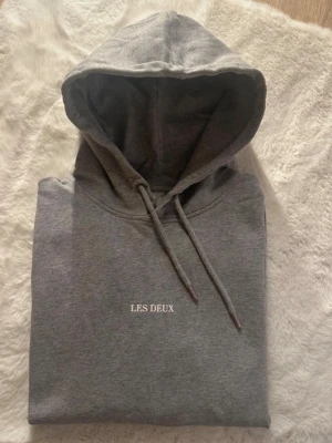 Grå hoodie från Les Deux - Snygg grå hoodie från Les Deux med minimalistisk vit logga på bröstet. Klassisk modell med huva och dragsnören samt en stor magficka framtill. Tillverkad i mjukt bomullsmaterial som känns skönt mot huden. Perfekt för en avslappnad och stilren look.