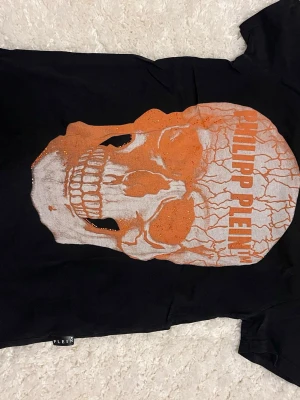 Svart Philipp Plein t-shirt med dödskalle - Cool svart t-shirt från Philipp Plein med stor orange och vit dödskallegrafik på framsidan och texten 'PHILIPP PLEIN'. T-shirten har korta ärmar och är tillverkad i mjuk bomull. Perfekt för dig som gillar edgy streetwear och vill sticka ut.