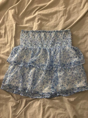 Blommig volangkjol i blått och vitt - Supersöt kort kjol med blått och vitt blommönster och volanger från veromoda . Kjolen har bred smock i midjan för skön passform och är gjord i ett lätt, luftigt material som passar perfekt till varma dagar. Perfekt att styla med en enkel topp eller hoodie.