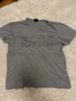 Grå Hugo Boss t-shirt med logga - Säljer en grå t-shirt från Hugo Boss med stor logga på bröstet. T-shirten har klassisk passform, rund halsringning och är kortärmad. Materialet känns mjukt och skönt, perfekt till vardags. Enkel och stilren design som funkar till det mesta.
