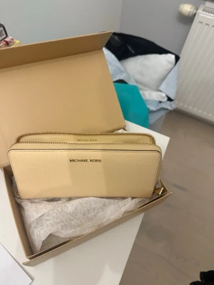 Beige plånbok från Michael Kors - Stilren beige plånbok från Michael Kors i saffianoläder med guldfärgade detaljer. Plånboken har dubbla dragkedjor, flera kortfack och ett fönster för ID eller kort. Perfekt för dig som vill ha ordning och snygg design i väskan.