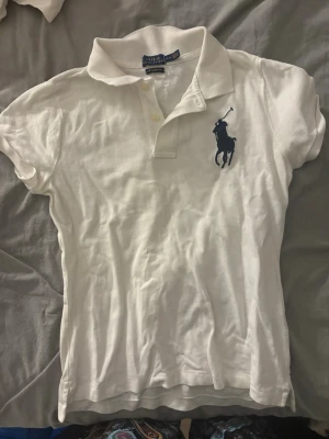 Vit pikétopp från Ralph Lauren - Klassisk vintage pikétopp från Ralph Lauren med korta ärmar och krage. Stort mörkblått Polo-logotryck på bröstet och knäppning framtill. Perfekt till jeans eller kjol för en sportig och stilren look.