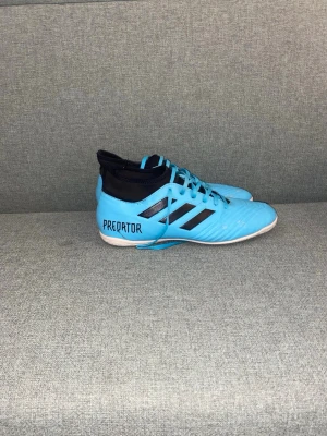 Adidas Predator blå inomhusfotbollsskor - Säljer ett par Adidas Predator inomhusfotbollsskor i blått med svarta detaljer och vita sulor. Skorna har snörning, högre ankelparti i svart och klassiska tre ränder på sidan. Perfekta för futsal eller inomhusträning.