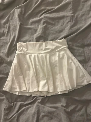 Vit plisserad skort med inbyggda shorts - Säljer en vit skort med plisserad kjoldel och bred midjeresår. Kjolen har inbyggda shorts under och är gjord i ett mjukt, stretchigt tyg som ger en sportig och trendig look. Perfekt för dig som vill ha både stil och komfort.