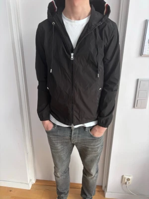 Moncler windbreaker - Snygg svart vindjacka från Moncler med dragkedja framtill, justerbar huva med dragsko och diskreta fickor på sidorna. Jackan har elastiska muddar vid ärmsluten och tunna ränder i rött och vitt på insidan av huvan. Perfekt för en stilren och sportig look.