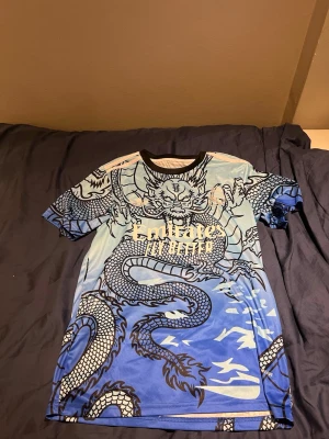 Blå Real Madrid tröja med drakmotiv - Säljer en blå Real Madrid matchtröja från Adidas med ett stort drakmotiv över hela tröjan. Framsidan har 'Emirates' tryckt och baksidan har 'Bellingham' och nummer 5. Tröjan har korta ärmar och svart krage. Perfekt för dig som älskar fotboll och unika designer.