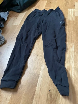 Svarta mjukisbyxor från Adidas - Säljer ett par svarta mjukisbyxor från Adidas med diskret logga på benet. Byxorna har resår i midjan, muddar vid bensluten och dragkedjeförsedda fickor. Perfekta för chill eller träning, tillverkade i mjukt material för maximal komfort.