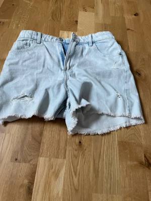 Ljusa denimshorts från H&M - Säljer ett par ljusa denimshorts från H&M med hög midja och avslappnad passform. Shortsen har råa, fransiga kanter och slitna detaljer framtill. Perfekta för varma dagar och en chill stil.