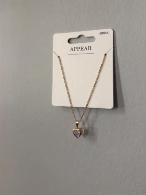 Guldhalsband med hjärthänge APPEAR - Snyggt guldigt halsband från APPEAR med ett hjärtformat hänge som har en glittrande sten i mitten. Kedjan är tunn och elegant, perfekt för att ge en söt touch till din outfit. Kommer på originalkortet.