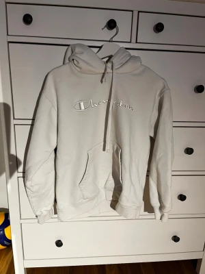 Vit hoodie från Champion - Säljer en stilren vit hoodie från Champion med broderad logga på bröstet. Hoodien har en stor känguruficka framtill, dragsko i huvan och ribbade muddar vid ärmar och nederkant. Perfekt för dig som gillar en clean och klassisk look.