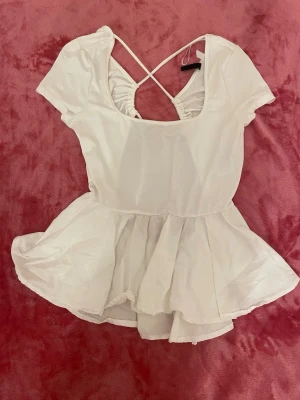 Vit body med kjol och korsade band - Söt vit topp med korta ärmar och volangkjol liknande nertill. Djup öppen rygg med korsade band och rynkade detaljer. Tillverkad i ett glansigt och mjukt material. Perfekt nu när det snart e vår!