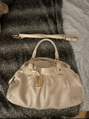 Beige handväska Marc by Marc Jacobs - Snygg beige handväska från Marc by Marc Jacobs i mjukt läder. Väskan har guldfärgade metalldetaljer, dubbla handtag och avtagbar axelrem. Märkets klassiska metallbricka framtill ger en exklusiv touch. Perfekt för dig som gillar stilrena och tidlösa accessoarer.
