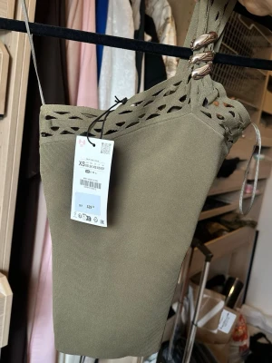 Olivgrön ribbad axelbandslös topp Zara - Trendig olivgrön ribbad topp med axelbandslös design och dekorativa metallringar på ena axeln. Toppen har ett snyggt hålmönster upptill och är croppad för en modern look. Perfekt att styla med höga jeans eller kjol. Ny med prislspp