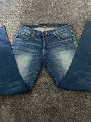 Blå Lee jeans - Säljer ett par blå jeans från Lee med raka ben och snygg slitning framtill. Sälj i typ ett otroligt bra skick. Perfekta för en avslappnad och trendig look. Hör av er vid fundering.