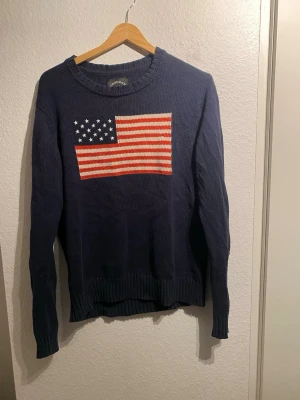 Stickad tröja från Crocker - Mörkblå stickad tröja med rund hals och ribbade muddar. På bröstet finns en stor amerikansk flagga i rött, vitt och blått. Tröjan har en avslappnad passform och är perfekt för dig som gillar klassisk, preppy stil. Går fint att pruta😁