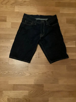 Mörkblå jeansshorts från Carhartt - Säljer ett par mörkblå jeansshorts från Carhartt med klassisk femficksdesign och kontrastsömmar. Shortsen har normal passform och ett Carhartt-märke på ena bakfickan. Perfekta för dig som gillar en enkel och tidlös stil.