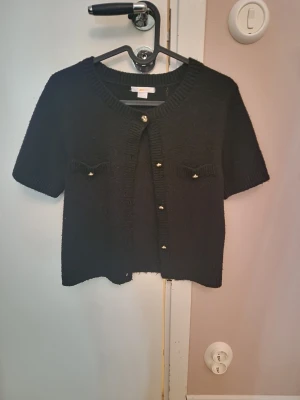 Svart kortärmad kofta med gulddetaljer - Snygg svart kofta med korta ärmar och ribbad kant vid halsen. Framtill finns dekorativa fickor och guldfärgade knappar som ger en lyxig touch. Koftan är stickad och har en croppad passform som passar perfekt till höga jeans eller kjol.