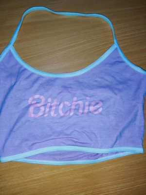 Lila croppad linnetopp med text bitchie  - Trendig lila croppad linnetopp med ljusblå kantband och texten 'Bitchie' i rosa framtill. Perfekt för dig som vill sticka ut och ha en edgy stil. Tunn och mjuk, troligen i bomull eller polyesterblandning. Axelbandslös design för en cool vibe. Små i storlek 
