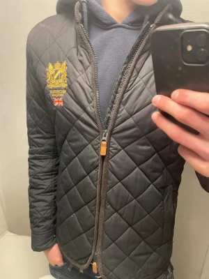 Svart quiltad jacka från Morris - Snygg svart quiltad jacka från Morris med gul broderad logga och brittisk flagga på bröstet. Jackan har dragkedja framtill, sidofickor och en klassisk, smal passform. Perfekt för dig som gillar stilrena detaljer och tidlös design.