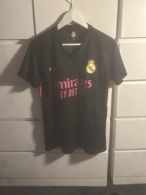 Real Madrid svart matchtröja Adidas - Snygg svart Real Madrid matchtröja från Adidas med klubbmärke på bröstet och rosa detaljer. Trycket 'Emirates Fly Better' på framsidan. Tillverkad i lätt och ventilerande material, perfekt för fotbollsträning eller sport.