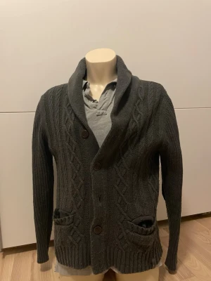 Mörkgrå kabelstickad kofta - Mysig mörkgrå kabelstickad kofta med sjalkrage och stora knappar framtill. Två fickor fram och ribbade muddar. Snygga detaljer med lappar på armbågarna. Perfekt för lager på lager och en avslappnad stil.