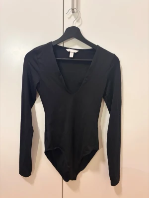 Svart långärmad body från H&M - En stilren svart body från H&M med lång ärm och djup V-ringning. Tillverkad i mjukt stretchigt material som sitter snyggt på kroppen. Perfekt att matcha med jeans eller kjol för en clean look.