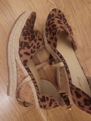 Leopardmönstrade espadrille-sandaler - Säljer ett par trendiga sandaler med leopardmönster och kilklack i kork och rep. Skorna har öppen tå, korsade band framtill och justerbar rem runt ankeln. Perfekta för dig som vill sticka ut med en djurisk look i sommar.