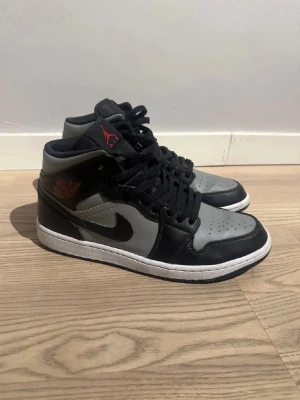 Air Jordan 1 Mid svart/grå - Säljer ett par Air Jordan 1 Mid sneakers i svart och grått med röda detaljer. Skorna har klassisk Nike Swoosh på sidan, svart snörning och vit sula. Tillverkade i läder och textil för en clean och tidlös look. Perfekta för dig som gillar streetwear och ikoniska sneakers.